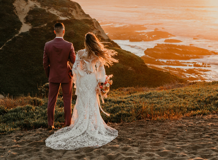 Groom and Bride standing on a grassy hill overlooking the ocean during sunset