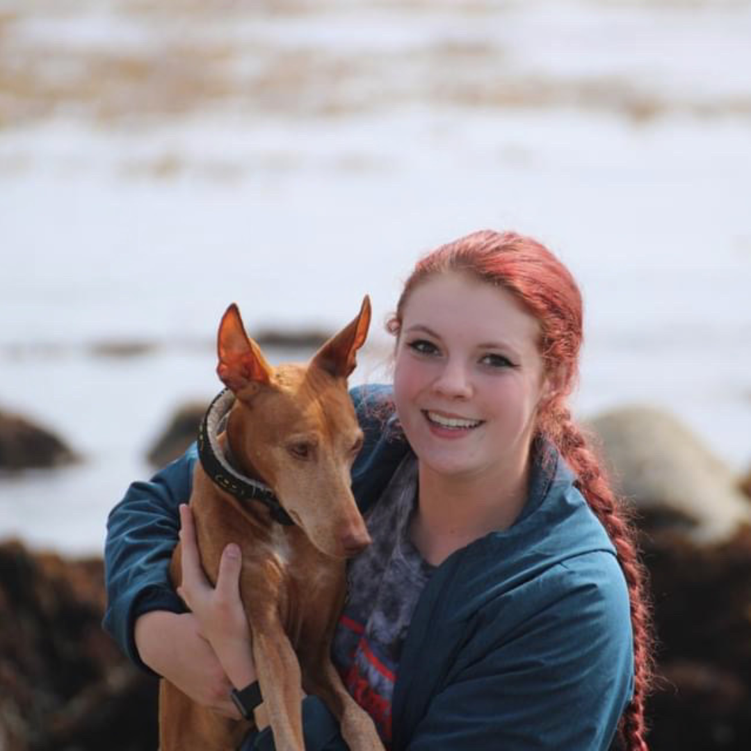 a woman with red hair hold a small dog. In front of the ocean